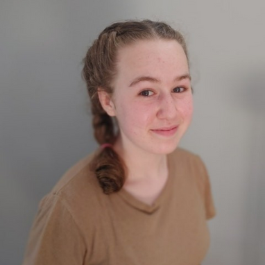 Young girl with light brown hair, in two French braids. She is wearing a light brown t-shirt.