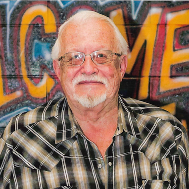 Older gentleman with white hair and beard. He is wearing glasses and a plaid shirt.