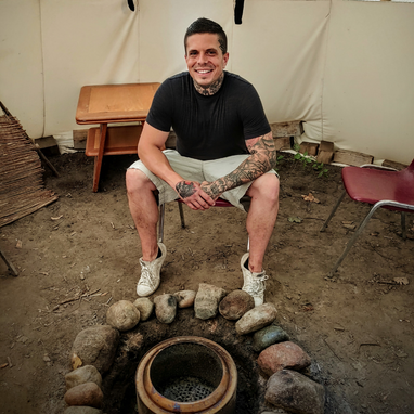 A man wearing a black t-shirt and light shorts. He has black hair and is sitting in front of a stone fire pit.