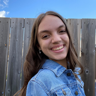 Young girl with light brown hair smiling. She is wearing a light blue jean jacket.